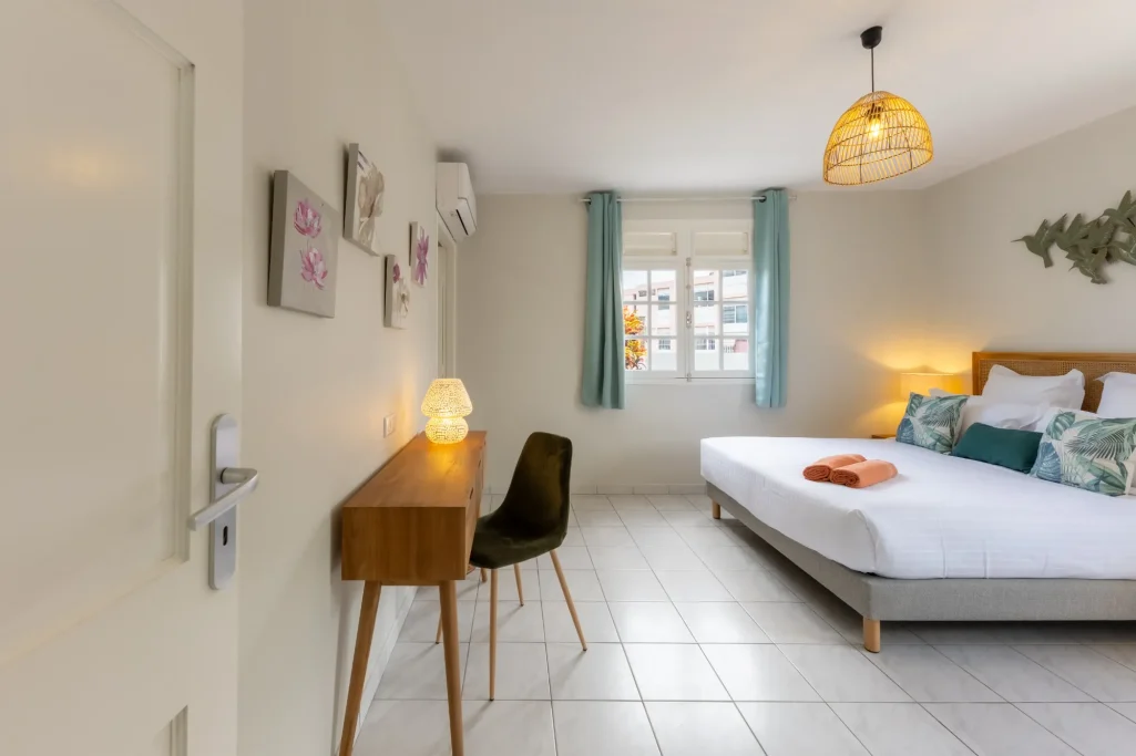 Chambre Garden Villa Bel Embrun Chambre lumineuse avec lit double, bureau, chaise et fenêtre aux rideaux turquoise.